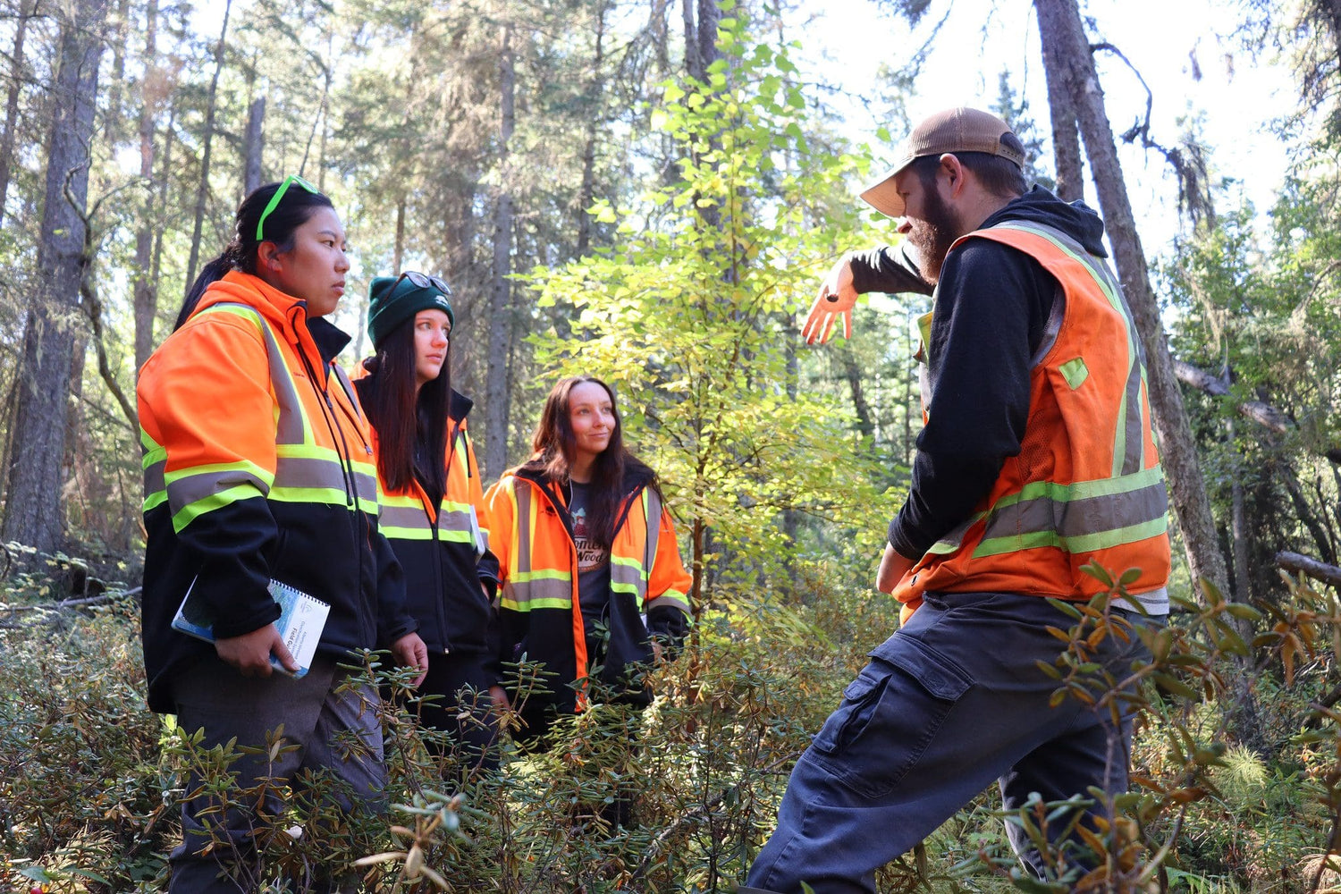 Group of people in safety vests in a forest setting with one person explaining something.